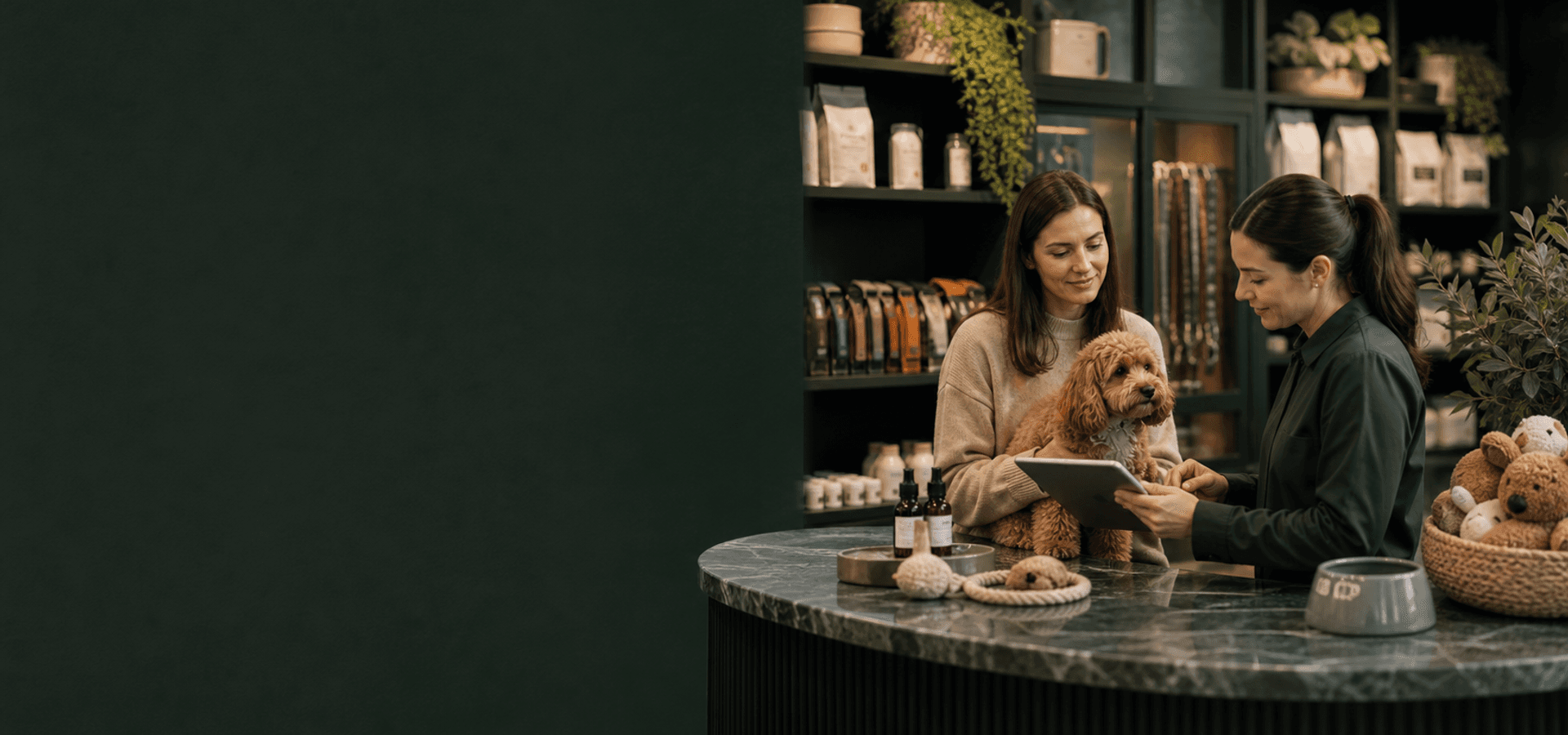 Profissional de pet shop em balcão moderno apresentando tablet de atendimento para tutora com cão calmo.